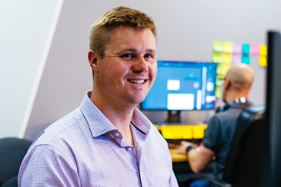 James, one of our developers, smiles for the camera as he is working, he is looking at his monitor. In the background we see the back of Luke who is working on his computer.