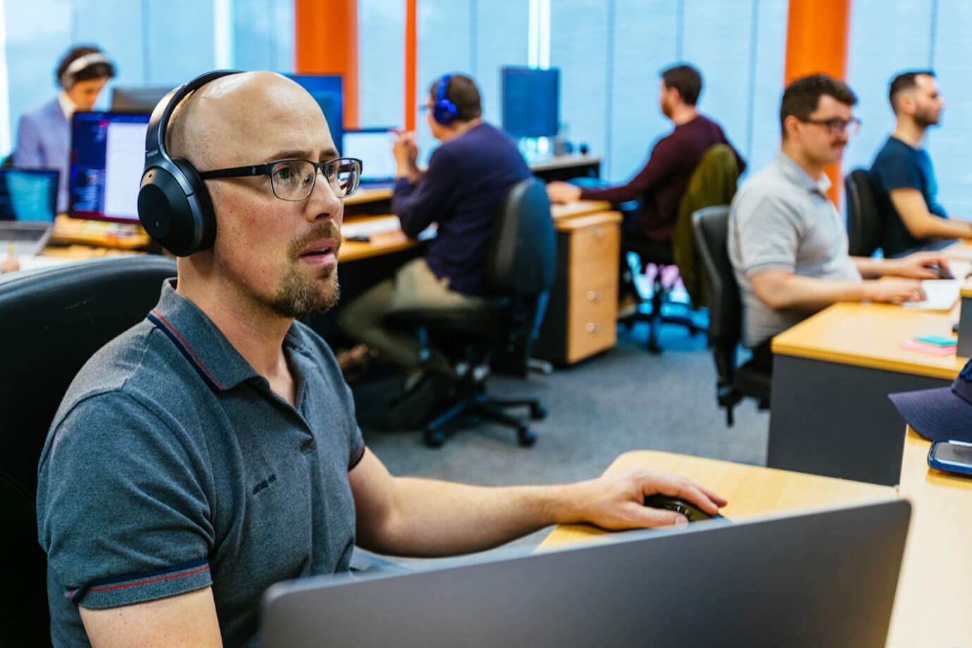 Luke, our UX and visual design specialist with headphones on, working. In the background, five Alliance developers are working.