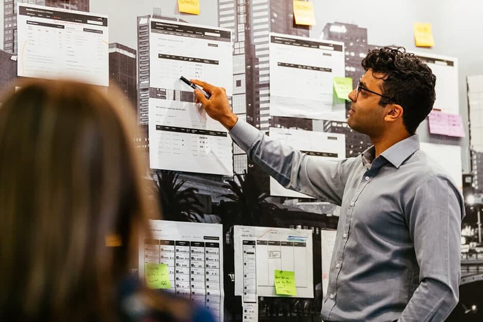 Naser, a project manager, who is pointing at printed wireframes stuck to the wall and discussing them with a client.