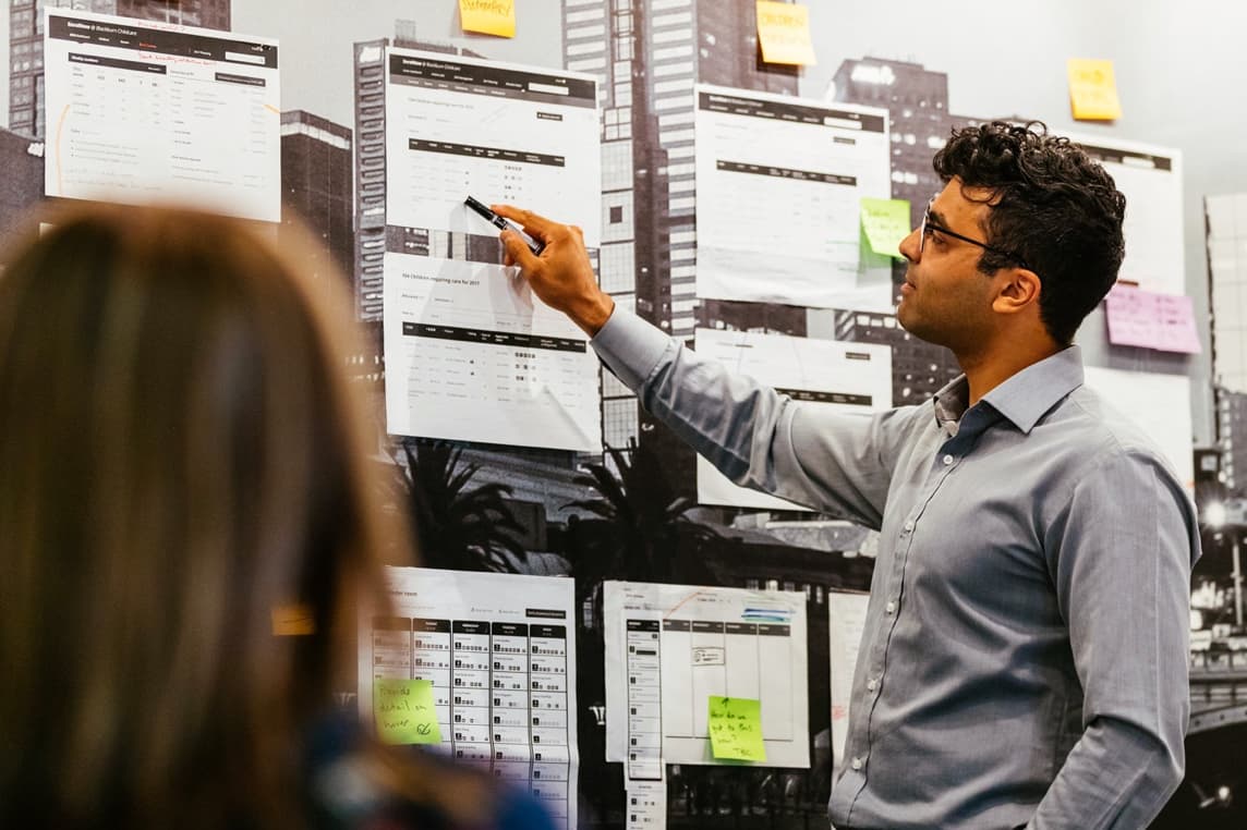 Naser, a project manager, who is pointing at printed wireframes stuck to the wall and discussing them with a client.